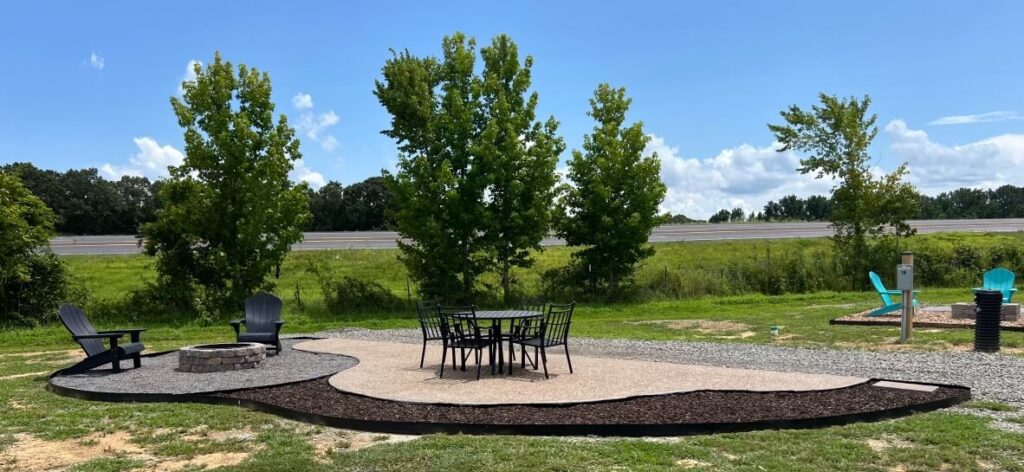 Outdoor patio area with a fire pit, Adirondack chairs, and a table with four chairs, surrounded by grass and trees, near a road in the background.